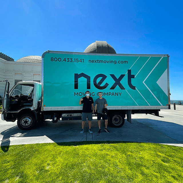 Two professional local movers standing at the front of the middle of the Next Moving box truck during the moving process. The truck is parked at the Griffith Observatory, Los Angeles, California. The body of the truck has Next Moving Company logo on it with the phone number 8004331541, and email address next.loc.